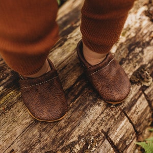 May include: A pair of brown leather baby moccasins with a textured finish. The moccasins are worn by a child standing on a log.