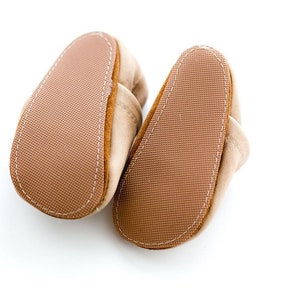 May include: A pair of brown suede baby booties with a textured sole. The booties have a white stitching detail around the edges.