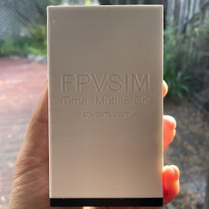 Puede incluir: Caja de plástico blanca con el texto "FPVSIM Timer Multi 8 Ch fpvsim.com" en relieve en la parte delantera. La caja tiene una base negra.