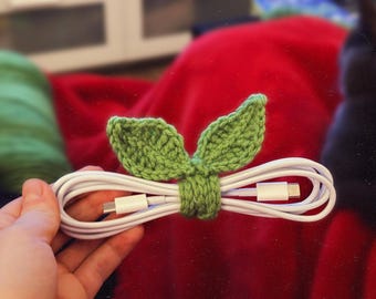 Puede incluir: Un cable de carga blanco se mantiene unido con un organizador de cable verde en forma de hoja de ganchillo. El organizador de cable está hecho de hilo verde y tiene dos formas de hojas. El fondo es una manta roja.