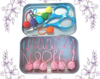 May include: A metal tin with a rainbow assortment of yarn loop stitch markers and pink stitch markers. The tin also contains a pair of small scissors.