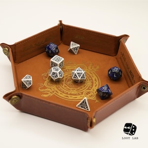 Puede incluir: Una bandeja de dados de cuero marrón con un diseño dorado y cierres de botón metálicos. La bandeja contiene varios dados poliédricos blancos y azules, perfectos para juegos de mesa.