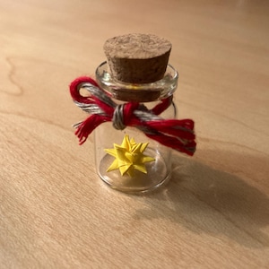 May include: A small glass jar with a cork lid and a red and white ribbon tied around the neck. Inside the jar is a yellow origami star.