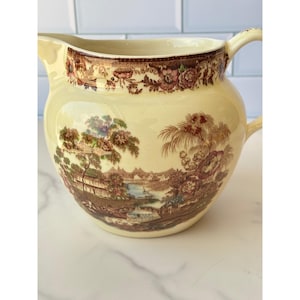 以下が含まれることがあります： 木々、川、建物が描かれた茶色の転写模様が特徴のクリーム色の陶器製ピッチャー。縁には花柄のボーダー模様が施されています。ピッチャーには曲線的なハンドルと、液体を注ぐための注ぎ口が付いています。