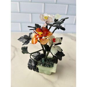 May include: A decorative bonsai-style sculpture featuring translucent carved stone flowers in shades of white, orange, and red with yellow and red centers. The plant has dark green leaves and is set in a pale green square stone planter.