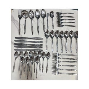 May include: A large set of stainless steel flatware featuring a floral pattern on the handles. The collection includes various forks, knives, spoons, a ladle, and a slotted spoon, all arranged in a grid on a white surface with gray veining.