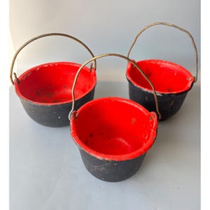 May include: Three small, cast iron cauldrons with red interiors and black exteriors. Each cauldron has a metal handle. The cauldrons are of varying sizes, with the largest approximately 6 inches in diameter.