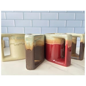 May include: A set of four ceramic mugs with a distinctive geometric square handle design. Each mug features a two-tone glaze with a cream-colored top section that drips into a solid base color of either off-white, dark brown, or deep red.
