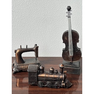 May include: A collection of three miniature metal figurines featuring a bronze-toned sewing machine, a violin on a stand, and a steam locomotive train. Each piece displays a weathered metallic finish with intricate molded details.
