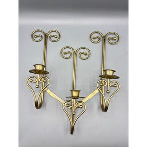 May include: A gold-toned metal wall sconce featuring three candle holders arranged in a tiered formation. Each holder is supported by a vertical rod topped with a scroll design and a heart-shaped base, connected by angled metal support bars.