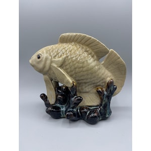 May include: A beige ceramic fish ornament with textured scales and fins, set on a dark brown and blue coral base. The fish has a glossy finish. The ornament is a decorative item, suitable for display. The background is plain.