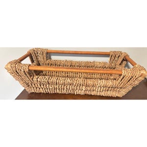 May include: Rectangular woven basket with wooden handles. The basket is made of natural-coloured woven material, with a rectangular shape and wooden handles on each end. The handles are a light brown colour. The basket is sitting on a brown surface.