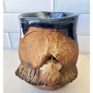 Könnte beinhalten: Ein handgefertigter Keramikbecher mit einem texturierten, erdbraunen Tonkörper und einem markanten, skulpturalen Gesicht. Der obere Rand ist mit einer glänzenden, dunkelblauen Glasur versehen, die über die raue, unglasierte braune Oberfläche läuft.
