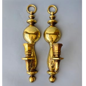May include: Pair of vintage brass wall sconces. Each sconce features a round top with a ring, a large spherical center, and a candle holder. The sconces have a polished, reflective surface and are mounted on a gray background.