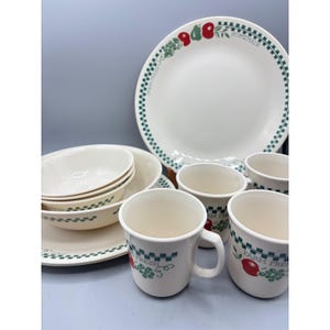 May include: A set of cream-colored ceramic dinnerware featuring a green checkered border and red fruit illustrations. The collection includes plates, bowls, and mugs, each decorated with the text "FARM FRESH" and "HOMEMADE" in a green script font.