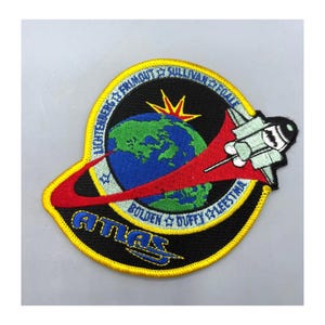 May include: Embroidered patch featuring a stylized space shuttle and a globe with a red arc. The patch has a black background with the word "ATLAS" and names around the edge. The patch is bordered with yellow and blue stitching.