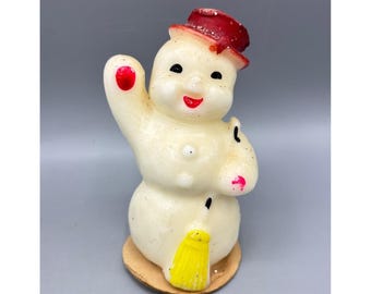 May include: A vintage snowman candle with a red top hat, black eyes, and a red smile. The snowman is holding up one arm with a red dot on the hand. It has a yellow broom and is standing on a brown base.