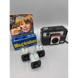 May include: A vintage black Magimatic camera with a flash cube system. The camera has the text "Magimatic" and "126" on it. The image also shows a box of Magicubes with the text "We make you smile!" and "12 Guaranteed Flashes".