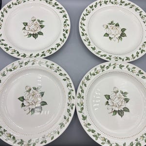 May include: Four white ceramic plates with a floral design. Each plate features a central white rose with green leaves and a border of green leaves and small white flowers. The plates have a diameter of approximately 25 cm.