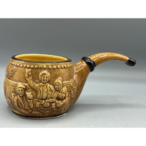May include: A brown ceramic pipe mug with a black rim and a raised design of three people drinking. The mug is shaped like a pipe with a curved handle.