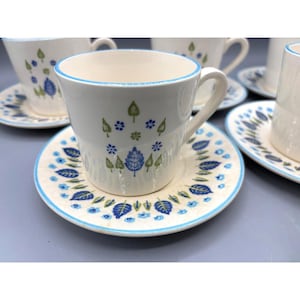 May include: White ceramic teacups and saucers with a blue and green leaf design. The teacups have a blue rim and handle. The saucers have a matching leaf pattern and blue trim. A set of vintage tableware.