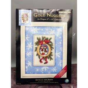 Puede incluir: Un kit de punto de cruz titulado "Gifts in the Snow" con un diseño enmarcado de dos muñecos de nieve dentro de un adorno dorado con un lazo rojo. El kit es una colección de 20 x 25 cm.