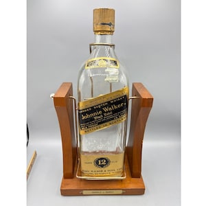 Puede incluir: Una botella de whisky escocés Johnnie Walker Black Label vintage en un soporte de exhibición de madera. La botella tiene una tapa dorada y una etiqueta con el texto "12 YEARS OLD" y "86.8 PROOF". El soporte de madera tiene una varilla de latón y una pequeña placa de identificación.
