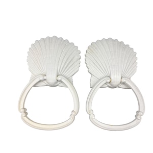 May include: Two white seashell-shaped drawer pulls with ring handles. The pulls have a detailed, textured design resembling a scallop shell. The ring handles are attached below the shell shape with decorative accents.