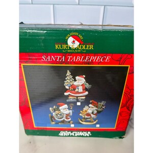 以下が含まれることがあります： Kurt S. AdlerのSanta Tablepiece用長方形の段ボール箱。緑と赤のパッケージには、Holidays Are Santa's World、Kurt S. Adler Since 1946、Santa Tablepiece、Snowtownという文字が印刷されており、3体のサンタクロースのフィギュアの画像が描かれています。
