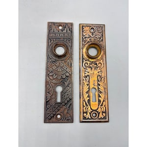 May include: Two antique door plates with intricate patterns. One is a dark bronze colour, the other is gold with a bronze keyhole. Both have a circular opening for the doorknob and are rectangular.