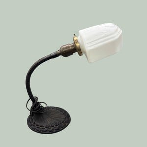 May include: A desk lamp featuring a round, dark metal base with a scalloped edge design, a flexible black gooseneck arm, and a brass-toned socket. The lamp is topped with a rectangular, white milk glass shade that has subtle molded geometric detailing.