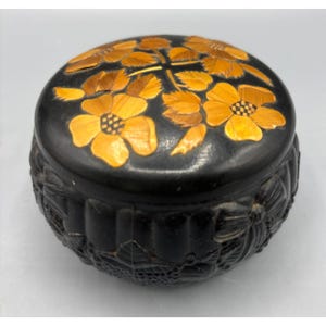 May include: A round, black decorative box with a lid. The lid is adorned with a floral design in shades of yellow and brown. The sides of the box feature a carved floral pattern, adding texture and detail to the piece.