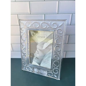 May include: A clear glass picture frame featuring an intricate embossed scrollwork pattern along the border. The center of the frame contains a display card with the text Marquis Waterford Crystal printed in black lettering.