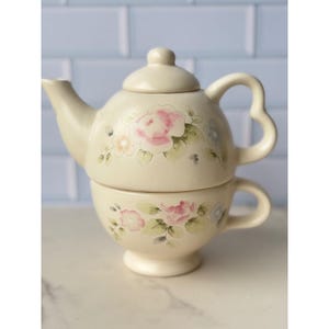 Puede incluir: Juego de té de cerámica color crema compuesto por una tetera y una taza. La tetera tiene forma redondeada con asa y boquilla curvadas. Ambas piezas están decoradas con diseños florales rosas y azules y hojas verdes.
