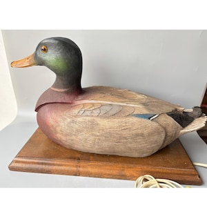 May include: A detailed, hand-painted duck figurine on a brown wooden base. The duck has a green head, brown body, and orange beak. The figurine is a decorative item, suitable for a home or office.