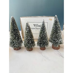 May include: A set of four miniature artificial pine trees with frosted white tips, each mounted on a small circular wooden base. The trees are arranged in front of a white cardboard box labelled Village Frosted Topiaries, Set of 4 Pieces.
