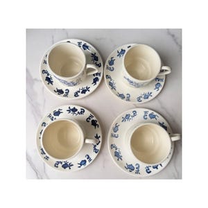 May include: Four cream-colored ceramic cups and matching saucers arranged in a grid. Each piece features a repeating pattern of dark blue botanical illustrations, including small fruits and leaves, encircling the rims and the cup exteriors.