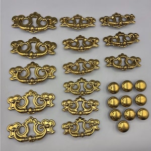 May include: A selection of vintage brass drawer pulls and knobs. The drawer pulls feature an ornate, scrolled design with a central handle. The knobs are round and solid. The hardware has a polished gold finish.