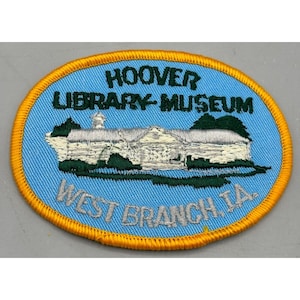 Puede incluir: Un parche bordado ovalado azul y amarillo con el texto "HOOVER LIBRARY-MUSEUM WEST BRANCH, IA". El parche presenta un edificio blanco con árboles verdes y un cielo azul.