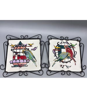 May include: Two square ceramic tiles with bird designs, each set in a decorative black metal frame. One tile features a bird in a cage, the other shows two birds. The birds are in shades of green, blue, and red.