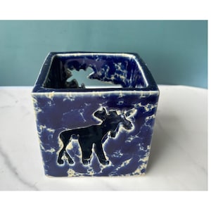 Può includere: Un portacandele quadrato in ceramica con una smaltatura blu profondo e un motivo bianco screziato. I lati sono ritagliati per rivelare la sagoma di un alce, creando un design traforato che permette alla luce di passare attraverso la struttura.