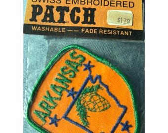 May include: A vintage orange embroidered patch with a green border. The patch features the outline of the state of Arkansas with a pine tree inside. The words "Arkansas" are embroidered in blue and white stars are embroidered around the state outline. The patch has a label that reads "Swiss Embroidered Patch Washable - Fade Resistant $1.79".