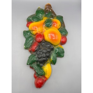 May include: A ceramic wall hanging featuring a cluster of fruit, including pears, grapes, and cherries. The fruit is painted in shades of yellow, red, green, and black, and the leaves are green.