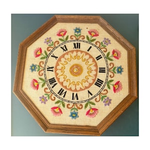 May include: An octagonal wall clock featuring a beige fabric face with hand-embroidered floral motifs in pink, blue, and purple. The clock face displays black Roman numerals and a central circular pattern in orange and yellow, all encased in a brown wooden frame.