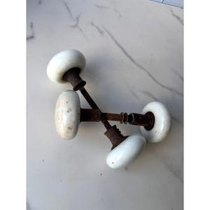 May include: Two vintage door knobs featuring smooth, round white porcelain heads attached to weathered, dark brown rusted metal spindles and collars. The knobs are positioned in an overlapping, crossed arrangement, showing signs of age and wear.