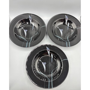May include: Set of three black ceramic soup bowls with a silver rim. Each bowl features a white and purple floral design with teal and gray accent lines. The bowls are round and have a deep center.
