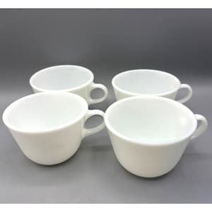 May include: Four white ceramic teacups with rounded bodies and attached handles. The cups are arranged in a square formation against a gray background. The cups are all the same size and shape.