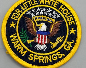 May include: A round, yellow-bordered patch with a black background. The patch features a white eagle with a red, white, and blue shield on its chest. The eagle is holding a green olive branch in its left talon and a bundle of wheat in its right talon. The text "FDR LITTLE WHITE HOUSE WARM SPRINGS, GA" is embroidered around the eagle in a yellow thread.