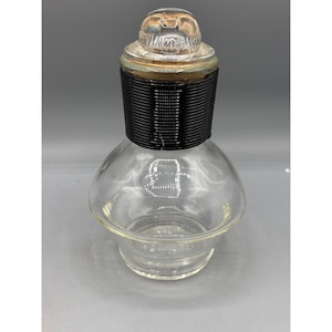 May include: A clear glass bottle with a black and silver lid. The bottle has a round body and a wide mouth. The lid has a textured design.