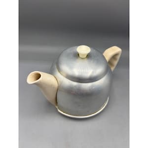 Puede incluir: Una tetera vintage de aluminio con asa, boquilla y pomo de tapa de color crema. La tetera tiene un cuerpo redondeado y una base pequeña y circular. La boquilla es curva y el asa está arqueada.